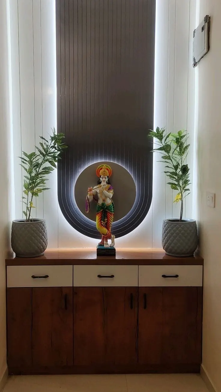 Pooja unit: a wood-finish cabinet with three drawers, below a backlit fluted panel with a round lit niche for the idol and a potted plant on each side